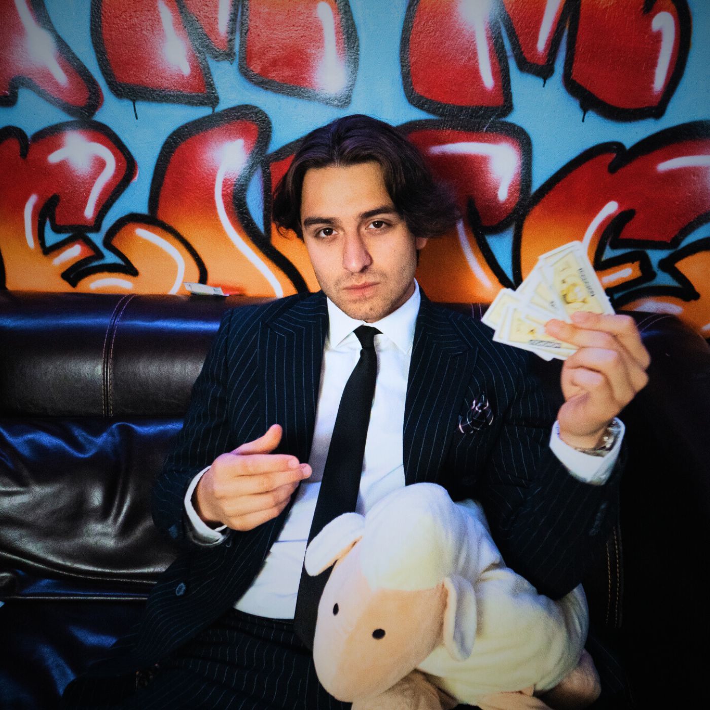 BaranBeatzz im Nadelstreifenanzug auf einem Ledersofa vor Graffiti-Wand. In der einen Hand hält er Spielgeld, in der anderen ein Plüschschaf – ein selbstironisches Coverbild zum Song „Besser Als Du“, das Luxus und Kindlichkeit pointiert gegenüberstellt.